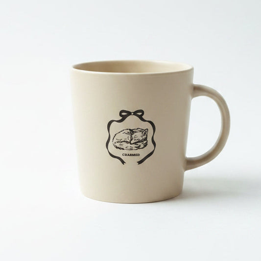 A cream-colored ceramic mug with a black line-art illustration of a sleeping cat inside a bow-shaped frame and the word 'CHARMED' below it